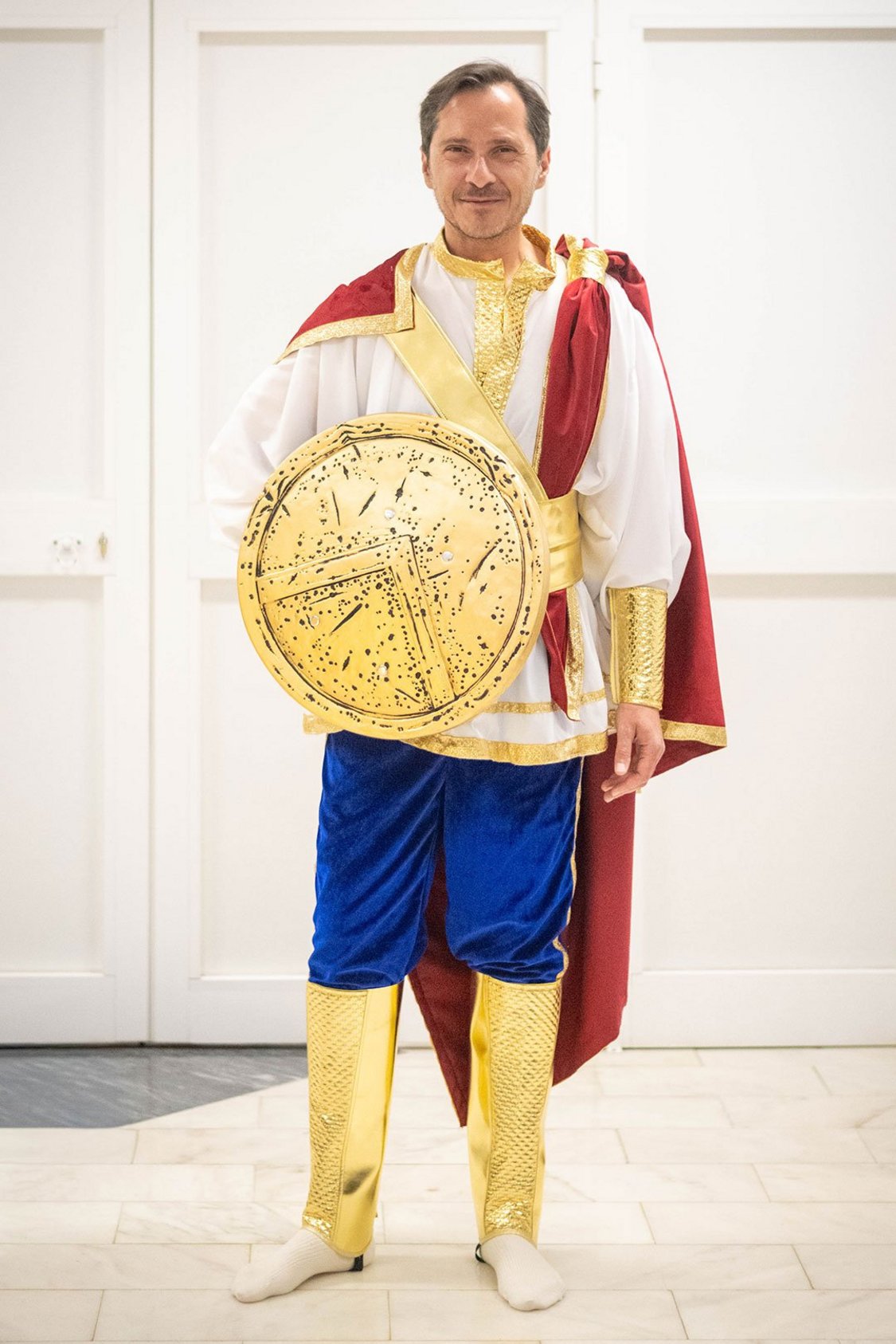 Figurino de Ulisses, com um escudo dourado, calça azul, sapato dourado, capa vermelha com detalhes em dourado e camisa branca
